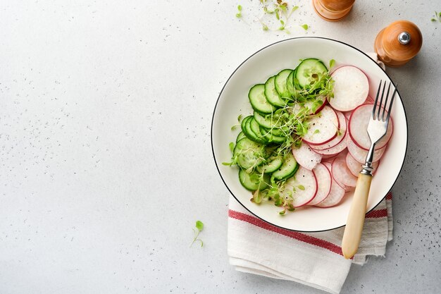 Frischer Salat mit rotem Rettich, Gurke, Gemüse, mikrogrünen Radieschen in weißer Platte auf grauem Steinhintergrund. Von oben betrachten. Konzept vegan und gesunde Ernährung.