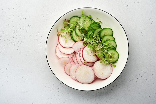 Frischer Salat mit rotem Rettich, Gurke, Gemüse, mikrogrünen Radieschen in weißer Platte auf grauem Steinhintergrund. Von oben betrachten. Konzept vegan und gesunde Ernährung.