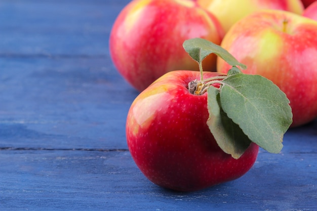 Frischer schöner roter Apfel mit grünen Blättern und vielen Äpfeln im Hintergrund auf einem blauen Holztisch