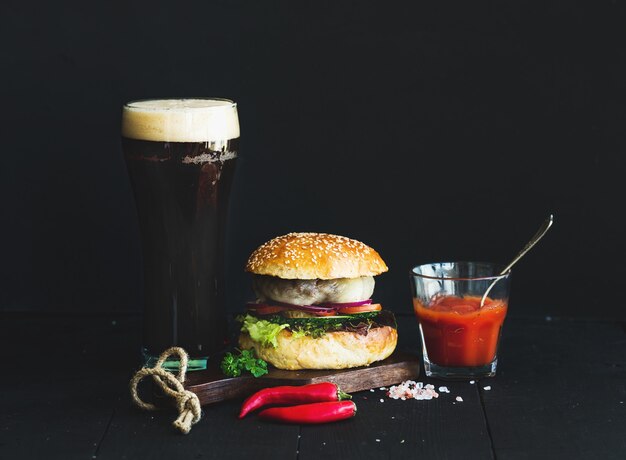 Frischer selbst gemachter Burger auf hölzernem Umhüllungsbrett mit würziger Tomatensauce, Seesalz, Kräutern und Glas dunklem Bier