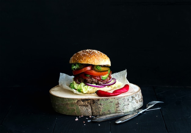 Frischer selbst gemachter Burger auf hölzernem Umhüllungsbrett mit würziger Tomatensauce, Seesalz und Kräutern über schwarzem Hintergrund.