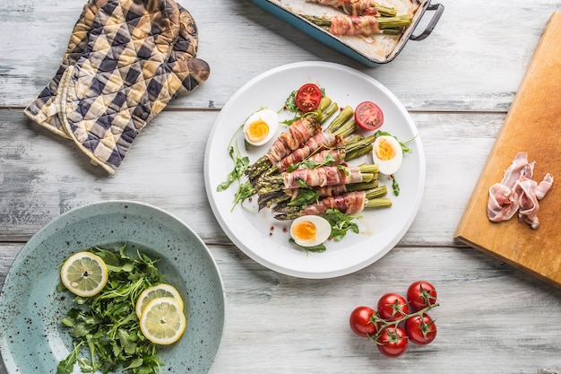 Frischer Spargel eingewickelt in Speck auf einem weißen Teller mit Rucola-Tomaten und Eiern von oben.