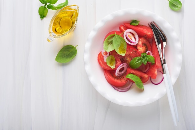 Frischer Tomatensalat mit Basilikumblättern, Olivenöl und Zwiebeln in weißer Schüssel auf hellem Hintergrund Traditionelle italienische oder mediterrane Ernährung Draufsicht