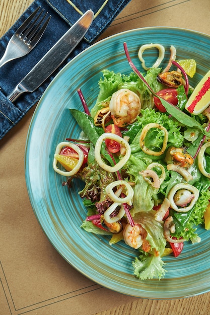 Frischer und grüner Salat mit Meeresfrüchten (Garnelen, Tintenfisch, Muscheln, Jakobsmuscheln), Kirschtomaten in einem blauen Teller auf einem Holztisch. Tischdekoration im Restaurant