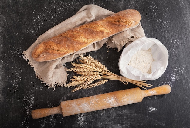 Frisches Brot mit Weizenährenmehl und Küchenutensilien auf dunklem Brett