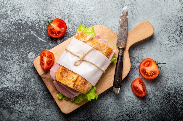 Frisches Ciabatta-Sandwich mit Schinken, Käse, Salat, Tomaten auf Holzbrett mit gealtertem Messer auf Steinbetonhintergrund, Nahaufnahme, Draufsicht. Gesundes Sandwich-Konzept machen