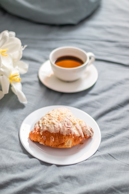 Frisches Croissant auf einer weißen Glasplatte und eine Tasse Kaffee auf dem Bett. Frühstück im Bett. Strauß weißer Tulpen.