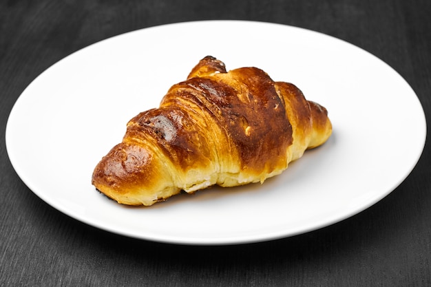 Frisches Croissant mit einer braunen Kruste auf einem weißen Teller auf einem schwarzen Tisch.