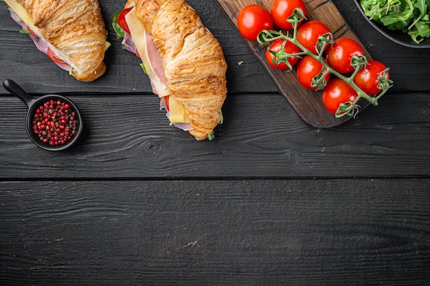 Frisches Croissant-Sandwich mit Schinken-, Käse- und Salatblattsatz, mit Kräutern und Zutaten, auf schwarzem Holztischhintergrund, Draufsicht flach, mit Kopierraum für Text