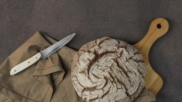 Frisches dunkles Brot auf dunklem Hintergrund durch Holzschüssel mit Vollkornmehl und Küchentuch in der Nähe