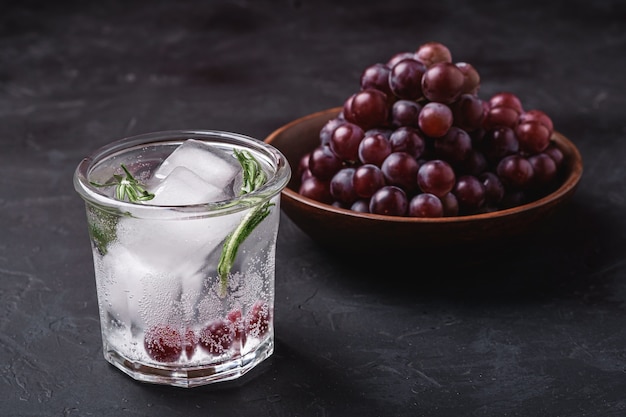 Frisches eiskaltes kohlensäurehaltiges Wasser im Glas mit Rosmarinblatt nahe der hölzernen Schüssel mit Traubenbeeren, dunklem Steinhintergrund, Winkelansicht