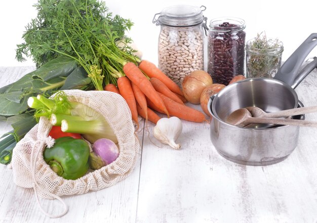 Frisches Gemüse in einem wiederverwendbaren Beutel und trockene Bohnen im Glas mit Zwiebeln, Knoblauch und Pfanne auf einem Tisch