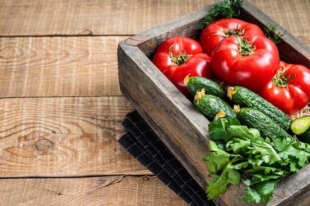 Frisches Gemüse in einer Holzkiste, rote Tomaten, grüne Gurken mit Kräutern. Hölzerner Hintergrund. Ansicht von oben. Platz kopieren.