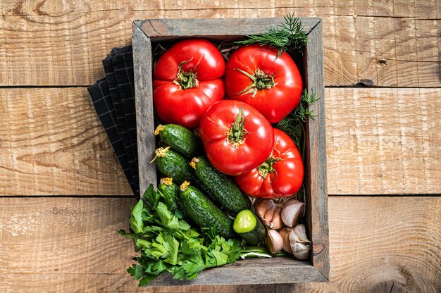 Frisches Gemüse in einer Holzkiste, rote Tomaten, grüne Gurken mit Kräutern. Hölzerner Hintergrund. Ansicht von oben.