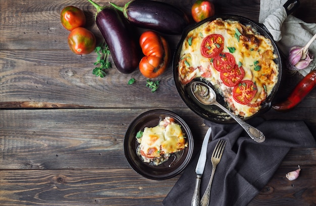 Frisches hausgemachtes Moussaka in Eisenpfanne auf rustikalem Holzhintergrund mit Zutaten griechischer Küche