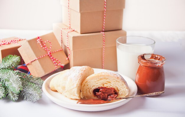 Frisches Hörnchenbrötchen mit Schokolade auf der Platte, dem Glas Milch, den Geschenkboxen und der Kiefer verzweigen sich auf den weißen Hintergrund