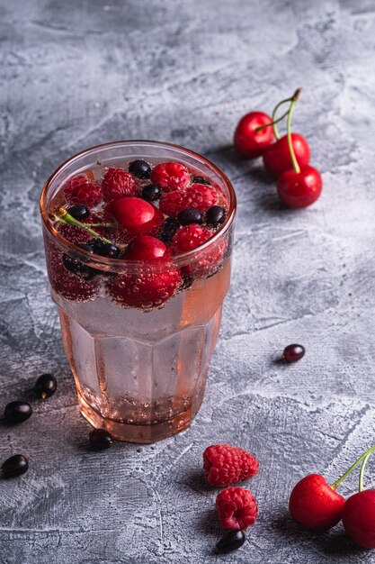 Frisches kaltes Sprudelwassergetränk mit Kirsch-, Himbeer- und Johannisbeerbeeren in rotem facettiertem Glas auf Steinbetonhintergrund, Sommerdiätgetränk, Winkelansicht