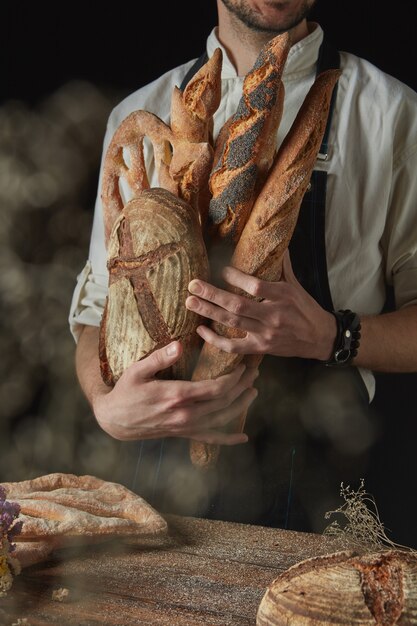 Frisches knuspriges Brot in den Händen eines Bäckers auf dunklem Hintergrund nahe einem Holztisch mit rundem Brot