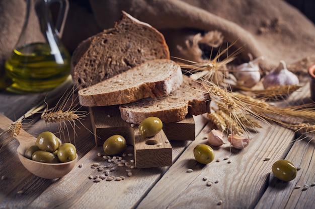 Frisches knuspriges Mehrkornbrot, grüne Oliven und Weizenähren auf einem rustikalen Holztisch. Bäckerei- und Lebensmittelgeschäft-Konzept.