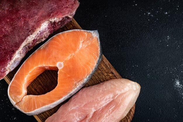 Frisches rohes Fleisch, Lachs und Hühnerfilet auf Holzbrett auf schwarzem Hintergrund. Natürliche Nahrung mit hohem Proteingehalt. Ansicht von oben.