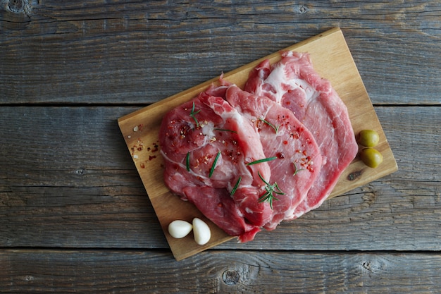 Frisches rohes Fleisch, Schweinesteaks in Gewürzen mit Oliven und Knoblauch auf einem Holzbrett auf einem alten rustikalen Holztisch