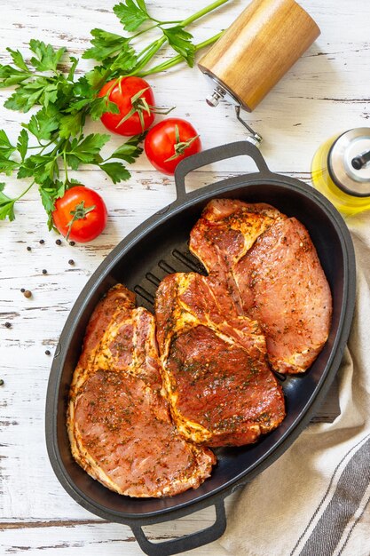 Frisches rohes Schweinelende am Knochen mit Gewürzen und Marinade in einer Gusseisenpfanne auf Holztisch Vorbereitung zum Backen Draufsicht flach gelegt