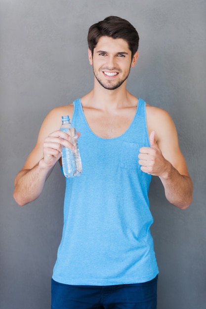 Frisches und sauberes Wasser nach gutem Training! Lächelnder junger muskulöser Mann, der eine Flasche mit Wasser und Daumen hoch hält, während er vor grauem Hintergrund steht