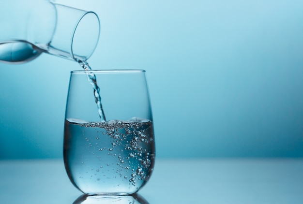 Foto frisches wasser, das vom krug in ein glas auf dem blauen hintergrund fließt. trinkwasser ist wichtig für eine optimale gesundheit