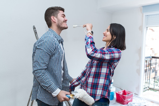 Frischvermählte haben Spaß an seiner neuen Wohnung während der Renovierung. Junge lateinische Frau, die mit ihrem Ehemann im neuen Zuhause mit Bürste spielt.