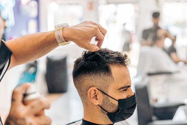 Friseur, der einem kaukasischen Mann, der eine Maske in einem Friseursalon trägt, Lack auf das Haar aufträgt