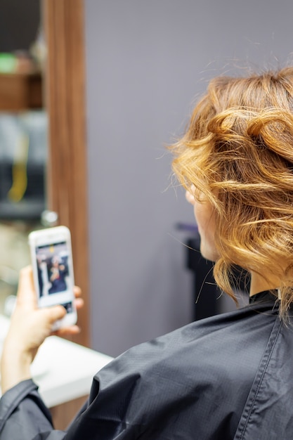 Friseur, der Frisur für junge Frau mit rotem lockigem Haar und mit Smartphone in ihren Händen im Schönheitssalon tut