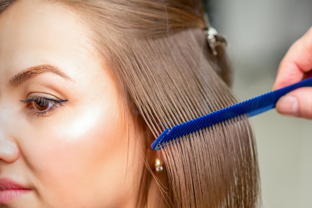 Friseur glättet weibliches braunes Haar mittlerer Länge mit einem Haar mit einem eisernen Haarglätter und einem Kamm in einem Schönheitssalon.