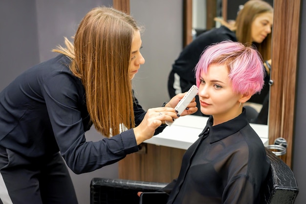 Friseur mit Kamm prüft und repariert die kurze rosa Frisur der jungen weißen Frau in einem Friseursalon