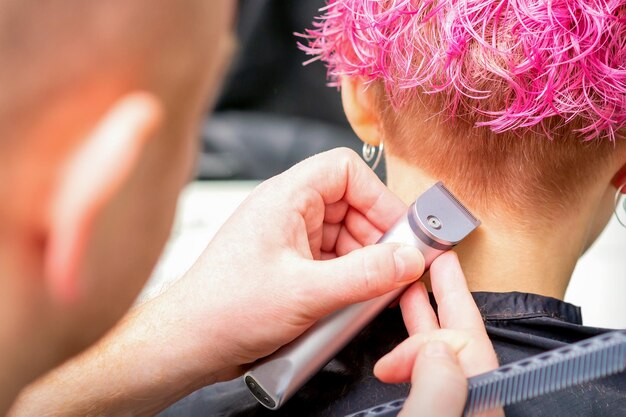 Friseur rasiert Nacken und Hals mit elektrischem Trimmer einer jungen kaukasischen Frau mit kurzen rosa Haaren in einem Schönheitssalon.