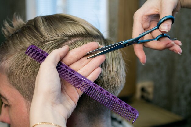 Friseur. Schließen Sie herauf die Hände der Frau, die den Haarschnitt des Mannes mit einer Schere und einem Kamm machen