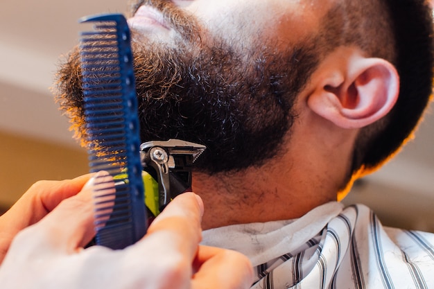 Friseur schneidet einen Bart mit einem Trimmer zu einem jungen hübschen Kerl mit einem Schnurrbart