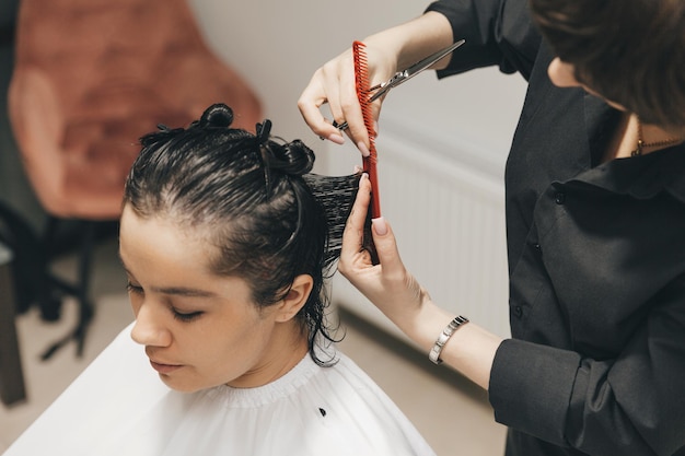 Friseur schneidet einer Frau im Salon die Haare. Der Friseur schneidet nasse Haare, indem er mit einem Kamm kämmt. Kunde mit kurzen Haaren, Rückansicht