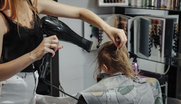 Friseurfrau macht modische hübsche Frisur für süße kleine blonde Mädchen im modernen Friseursalon, Friseursalon. Friseur macht Frisur für junges Baby im Friseurladen. Konzept Frisur und Schönheit