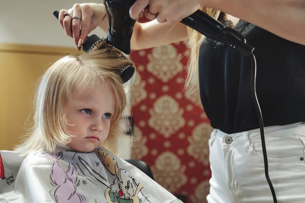 Friseurfrau macht modische hübsche Frisur für süße kleine blonde Mädchen im modernen Friseursalon, Friseursalon. Friseur macht Frisur für junges Baby im Friseurladen. Konzept Frisur und Schönheit