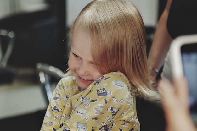 Friseurfrau macht modische hübsche Frisur für süße kleine blonde Mädchen im modernen Friseursalon, Friseursalon. Friseur macht Frisur für junges Baby im Friseurladen. Konzept Frisur und Schönheit