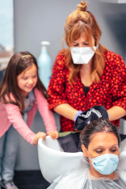 Friseurin mit Maske und Handschuhen wäscht die Haare der Kundin und ihrer Tochter, die geflügelt spielt