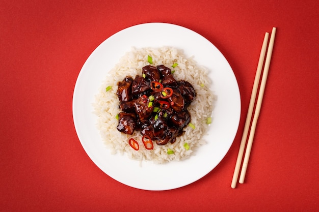 Frite a carne agridoce em molho pegajoso com arroz na chapa branca com pauzinhos de madeira, prato tradicional chinês. Jantar asiático ou almoço em fundo vermelho, close-up, vista de cima