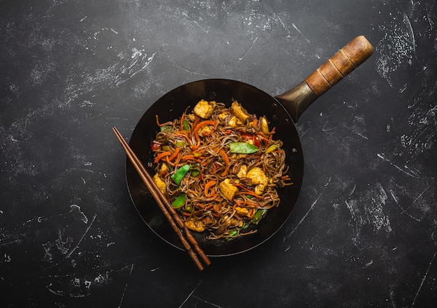 Frite o macarrão soba com frango, legumes na velha frigideira wok rústica, pauzinhos no fundo de pedra preta, close-up, vista superior. Refeição tradicional asiática / tailandesa, close-up