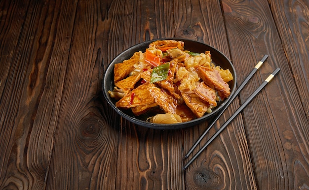 Frite tofu caseiro com legumes em molho doce quente e um par de pauzinhos na mesa de madeira escura e copie o espaço. Vista superior do conceito de comida asiática