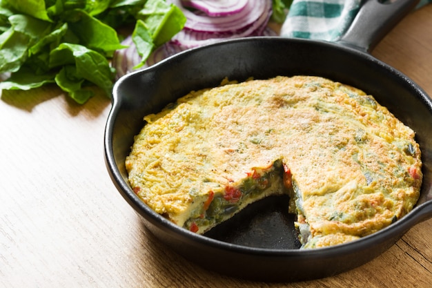 Frittata hecha de huevos y verduras en una sartén de hierro, sobre una mesa de madera.