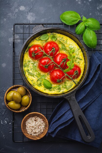 Frittata mit Rucola, Kartoffeln und Kirschtomaten in Eisenpfanne auf altem Steintisch. Ansicht von oben.