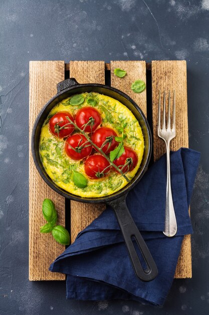 Frittata mit Rucola, Kartoffeln und Kirschtomaten in Eisenpfanne auf altem Steintisch. Ansicht von oben.