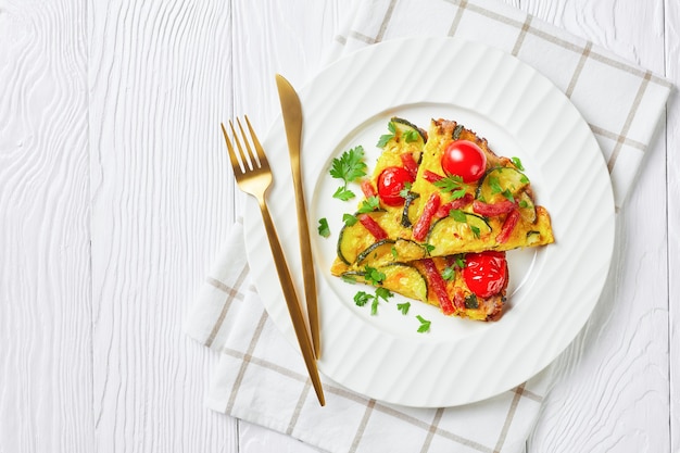 Frittata, tortilla con calabacín, finas salchichas ahumadas y tomate relleno en un plato blanco sobre mesa de madera, vista horizontal desde arriba, plano, espacio libre