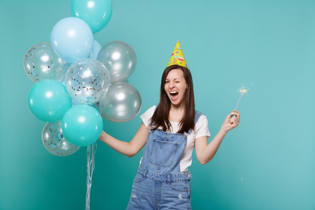 Fröhlich blinkende junge Frau mit Geburtstagshut, die eine brennende Wunderkerze hält und mit bunten Luftballons feiert, die auf blauem türkisfarbenem Hintergrund isoliert sind. Geburtstagsfeier, Menschen Emotionen Konzept.