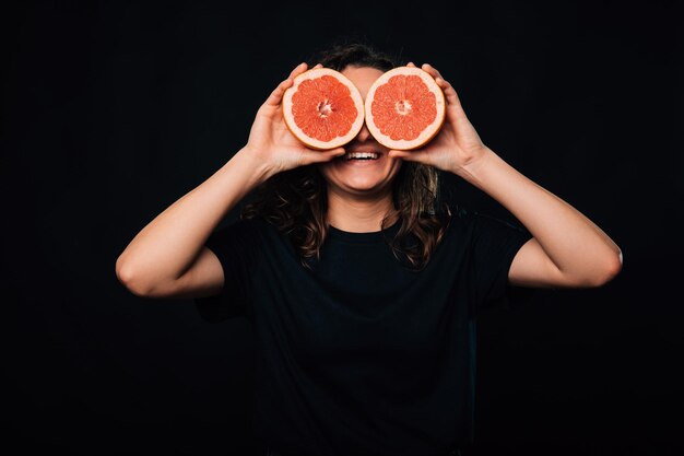 Fröhlich lächelnde Frau bedeckt ihre Augen mit zwei Hälften rosa Grapefruit